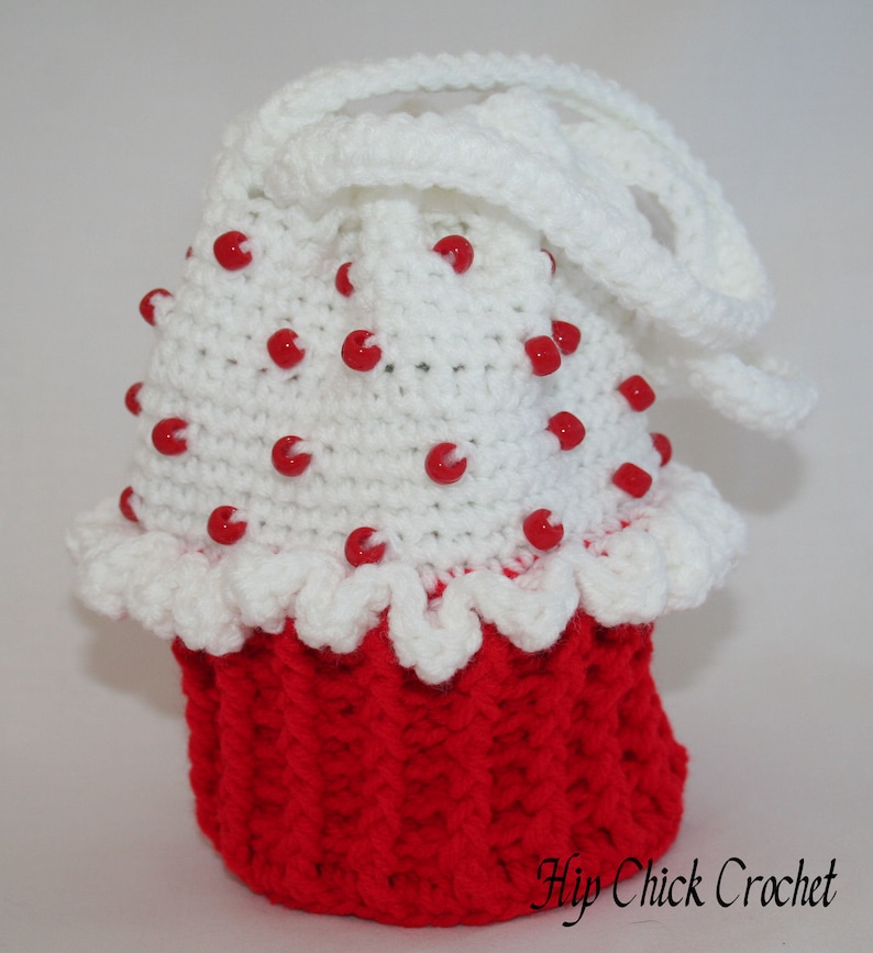 Peut inclure: Un sac en crochet blanc en forme de cupcake avec des perles rouges et une anse en crochet blanc. Le sac est rouge en bas et a un volant en crochet blanc autour du haut. Le texte "Hip Chick Crochet" est visible dans le coin inf&eacute;rieur droit de l'image.