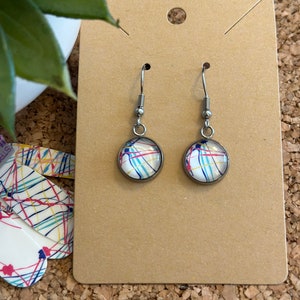 May include: A pair of silver dangle earrings with round glass cabochons featuring a colourful abstract design. The cabochons have a white background with red, blue, yellow, and black lines.