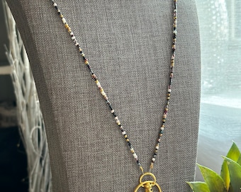 Pode incluir: Um cordão de contas preto, branco e dourado com um fecho giratório dourado. O cordão é apresentado em um manequim de tecido cinza.
