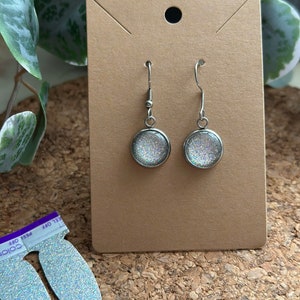 May include: A pair of silver dangle earrings with round, iridescent glitter-filled glass charms. The earrings are displayed on a brown card.