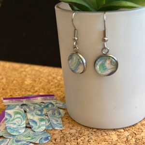 May include: A pair of silver dangle earrings with round blue and white marbled glass cabochons. The earrings are hanging from a white ceramic cup.