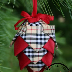 May include: A red and black plaid fabric Christmas ornament shaped like a pine cone. The ornament is hanging from a red ribbon and has a white circle with the text "Hip Chicks Digital File Design" on it.