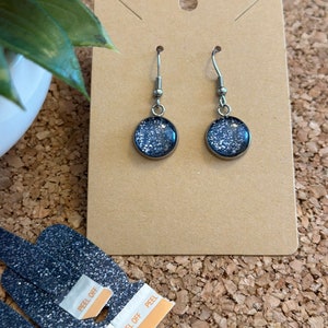 May include: A pair of silver dangle earrings with round, blue glitter-filled resin cabochons. The earrings are displayed on a brown card.