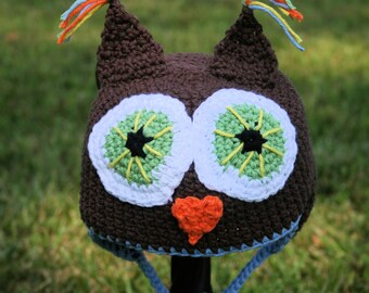 May include: A brown crocheted owl hat with green and white eyes and an orange felt beak. The hat has blue and white yarn ties.