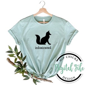 Op de afbeelding: Een lichtblauw T-shirt met een zwart silhouet van een vos en de tekst "infocxicated". Het T-shirt hangt aan een hanger en er is een groene tak op de achtergrond. De afbeelding bevat ook een rond logo met de tekst "Hip Chicks Digital File Design".