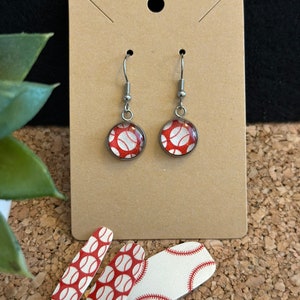 May include: Dangle earrings with a baseball theme. Each earring has a round design of a red and white baseball, set in a silver-tone metal frame. The earrings are displayed on a brown card, with baseball nail art strips below.
