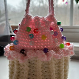 Peut inclure: Un sac en crochet en forme de cupcake rose et blanc avec des perles color&eacute;es autour du bord sup&eacute;rieur.