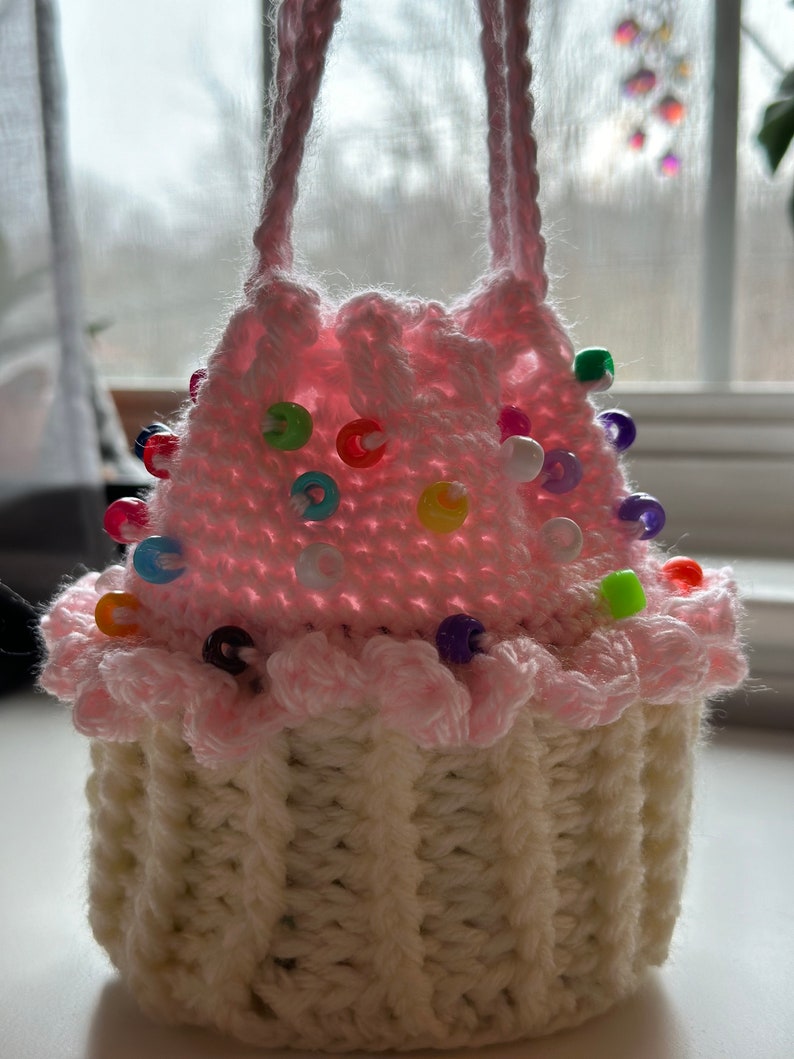Peut inclure: Un sac en crochet en forme de cupcake rose et blanc avec des perles color&eacute;es autour du bord sup&eacute;rieur.