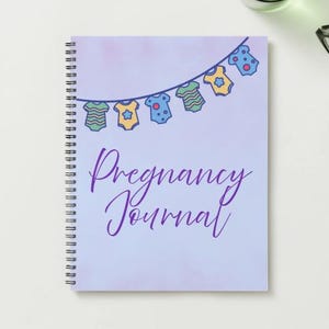 May include: A spiral-bound notebook with a light purple cover featuring the words "Pregnancy Journal" in cursive. The cover is decorated with a garland of baby clothes in blue, yellow, and green. The notebook is on a white surface.