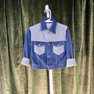 May include: A blue denim jacket with white and blue striped accents on the upper chest and cuffs. The jacket has two front pockets and gold-colored buttons. The jacket is hanging on a white hanger against a green curtain.