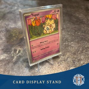 May include: A clear plastic card display stand holding a Togepi Pokémon card. The card features a cartoon illustration of Togepi with a floral background. The text "CARD DISPLAY STAND" is at the bottom.