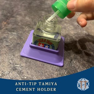 May include: A purple anti-tip Tamiya cement holder. The holder is designed to cradle a small bottle of Tamiya Extra Thin Cement. The bottle is clear with a green cap and a white applicator. The text "ANTI-TIP TAMIYA CEMENT HOLDER" is at the bottom.