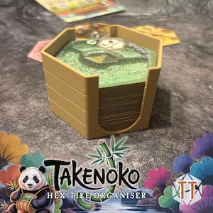 May include: A tan hex tile organizer for the board game Takenoko. The organizer holds a stack of game tiles with a green landscape design. The Takenoko logo and a panda graphic are visible.
