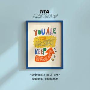 May include: A framed print with a blue border displays the words "YOU ARE THE PROJECT KEEP UPGRADING" in colourful, handwritten lettering. The print is on a light beige background and includes an orange arrow and stars. The words "TITA ART SHOP" are at the top.