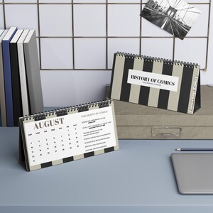May include: Desk calendar with black and beige vertical stripes, titled "History of Comics." The calendar is open to August, with a list of comic-related events. A small photo of the Brooklyn Bridge is pinned to the wall behind the calendar.