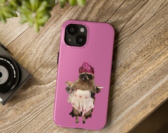 May include: A pink phone case featuring a raccoon wearing a pink crown and a floral apron, holding a martini and a cigarette. The phone case has a dual-lens camera and is set on a wooden surface.