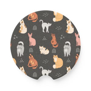 May include: A round, dark gray car coaster featuring a repeating pattern of cartoon cats in various colors and poses. The cats are white, orange, gray, and pink, with small rainbow and dash designs. The coaster has a small notch for easy removal.