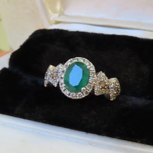 May include: An emerald-cut green gemstone ring with a halo of small diamonds. The ring has a gold band with additional diamond accents on each side. The ring is displayed on a black velvet surface inside a jewelry box.
