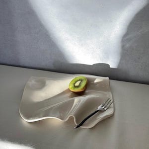 May include: A white, wavy-edged ceramic plate holds a halved kiwi fruit and a black-handled fork. The plate's design resembles draped fabric. The kiwi's green flesh and black seeds contrast with the plate's neutral color.