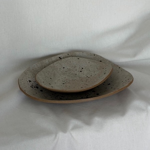 May include: Two speckled ceramic plates, one nested inside the other. The plates are light gray with dark speckles and a natural, unglazed rim. The larger plate has an organic, irregular shape.