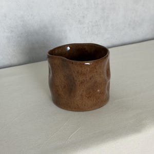 Puede incluir: Una pequeña taza de cerámica marrón hecha a mano con una superficie texturizada. La taza tiene una forma ligeramente irregular y un acabado liso y brillante. El interior de la taza es de color marrón oscuro y el exterior tiene un aspecto moteado.
