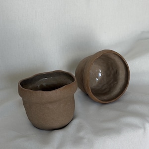 May include: Two small, handmade brown ceramic cups. One cup is upright, and the other is on its side. The cups have a rough, textured exterior and a smooth interior. The cups are on a white cloth background.
