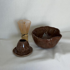 May include: A brown ceramic matcha bowl with an irregular, wavy rim, a matching brown ceramic whisk holder, and a light-coloured bamboo matcha whisk. The pieces feature a glossy, dark brown glaze with visible texture and organic, hand-formed shapes.