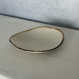 Puede incluir: Plato ovalado de cerámica blanquecina con borde marrón oscuro. El plato tiene una superficie lisa y una forma ligeramente irregular. El borde tiene un aspecto texturizado y pintado a mano. Adecuado para servir aperitivos o porciones pequeñas.