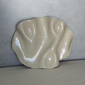 May include: A cream-colored, ceramic decorative plate with a wavy, organic shape. The plate has a glossy finish and is displayed on a neutral surface against a gray background. The design resembles a seashell.
