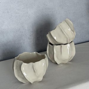 May include: Three off-white ceramic bowls with a unique, petal-like design. The bowls are stacked and arranged on a white surface, with a light gray background. The bowls have a glossy finish and a textured, organic shape.