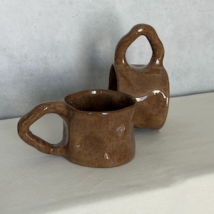 May include: A set of three ceramic mugs with a speckled, matte finish. The mugs are in shades of cream, light brown, and dark brown. Each mug has a simple, minimalist design and a rounded handle.