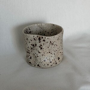 May include: A speckled ceramic cup with a cylindrical shape. The cup is off-white with dark brown speckles. The rim of the cup has an uneven, organic shape. The cup is likely handmade and suitable for beverages.