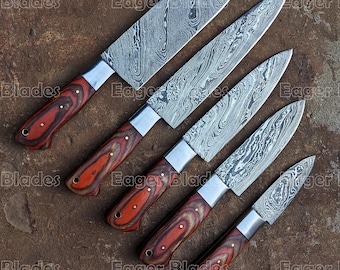 May include: Set of five kitchen knives with Damascus steel blades and wooden handles. The knives are arranged in a row from largest to smallest, with the largest knife at the top of the image. The knives have a distinctive pattern on the blades, which is characteristic of Damascus steel.