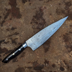 Może przedstawiać: Zbliżenie srebrnego noża szefa kuchni z ciemnym drewnianym uchwytem. Nóż ma falistą, wzorzystą klingę i leży na brązowej, fakturowanej powierzchni.