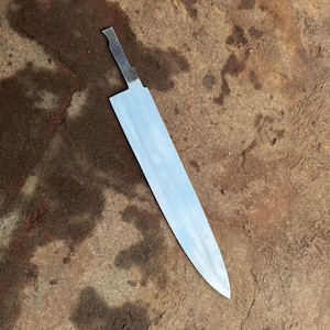 May include: A shiny, unfinished stainless steel knife blade with a long, straight edge and a pointed tip. The blade is lying on a rough, brown surface, showcasing its polished surface and potential for custom handles.