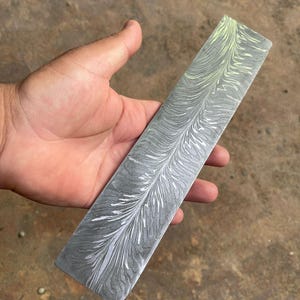May include: A rectangular metal billet featuring a dark gray base with a prominent white and pale yellow feather-like pattern running along its length. The surface displays a complex, swirling marbled texture characteristic of Damascus steel.