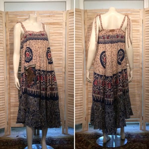May include: A long, flowing, beige dress with blue and red patterned fabric. The dress has a square neckline and thin straps. The dress has a tiered skirt with a ruffled hem.