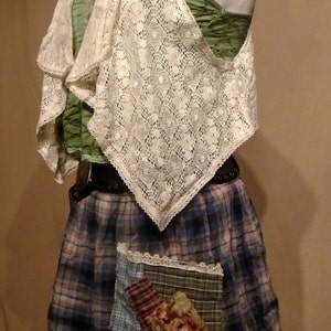 Può includere: Un gilet di pizzo bianco con un top verde sotto. Il gilet ha un orlo a festoni e viene indossato sopra una gonna a quadri con una stampa floreale sotto. La gonna ha un bordo in pizzo e un design patchwork.