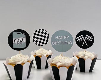 May include: Four cupcakes with white frosting and decorative toppers. The toppers include a fuel can, checkered flag, checkered circle, and a Happy Birthday message. The cupcake wrappers are black and white striped.
