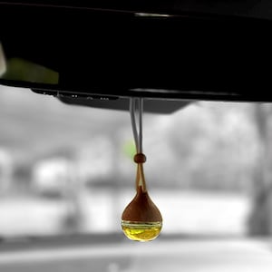 May include: A car air freshener hanging from a rearview mirror. The air freshener has a wooden top and a glass container filled with yellow liquid. The background is blurred, with a black rearview mirror and a grayscale view of the outside.
