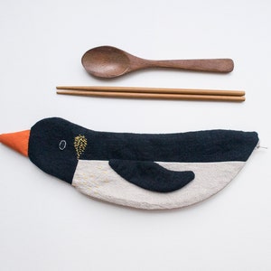 Puede incluir: Un porta cubiertos de tela con forma de pingüino en blanco y negro con cierre de cremallera. El porta cubiertos está sobre una superficie blanca con una cuchara de madera y palillos a su lado.