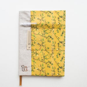 Könnte beinhalten: Ein gelber Stoff-Notizbuchumschlag mit einem Blumenmuster und einem kleinen gestickten Eichhörnchen. Der Umschlag hat eine Tasche auf der Vorderseite und einen Stifthalter oben.