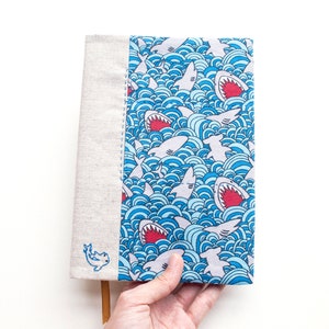 Op de afbeelding: Een boekomslag met een blauw en wit haaienpatroon. De omslag heeft een herhalend ontwerp van haaien met open monden in een golvende oceaan. De rug is van lichtbeige linnen met een kleine geborduurde haai.