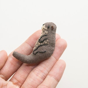 May include: A small, brown, hand-stitched stuffed otter with black eyes and a brown nose. The otter is sitting on a person's hand.