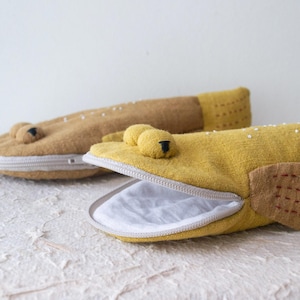 May include: Two fabric pencil cases shaped like fish, one in mustard yellow and one in tan. Each case features a white zipper mouth, protruding round eyes, and side fins with stitched details. The interior of the open yellow fish case is white.