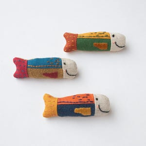 Può includere: Tre colorati giocattoli di peluche fatti a mano a forma di pesce. Ogni pesce ha una faccia sorridente ed è realizzato in tessuto di vari colori, tra cui blu, arancione, verde e giallo. I pesci sono lunghi circa 10 cm.