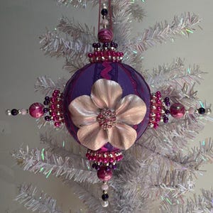 Puede incluir: Un adorno navideño esférico de color morado con una gran flor de color rosa pálido metálico en el centro con un racimo de pedrería. El adorno está decorado con cuentas rosas y moradas, detalles plateados y un lazo para colgar.