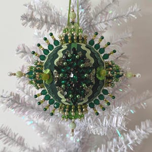 May include: A spherical ornament featuring a dark green fabric base adorned with an intricate arrangement of emerald green crystals, small white pearls, and gold-toned beads. The surface is decorated with wavy light green ribbon and beaded spikes.
