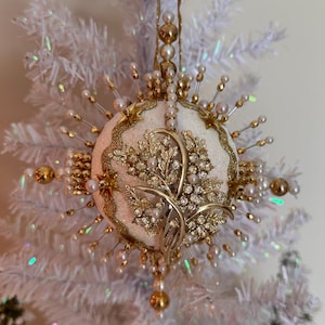 Puede incluir: Un adorno navideño esférico con una superficie blanca brillante decorada con un broche floral dorado, pequeñas perlas y detalles de estrellas metálicas. El adorno está adornado con alfileres radiantes rematados con perlas y cuentas doradas.