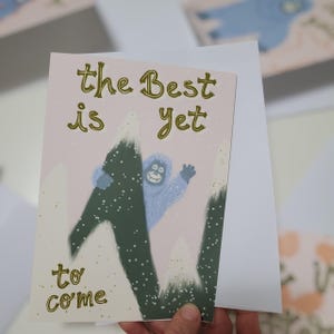 May include: A greeting card with the words "the Best is Yet to Come" in gold. It shows a light blue yeti waving from behind a green mountain with white snow. The background is a pale pink.